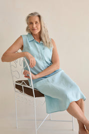 Woman wearing the Samara Linen Dress Sea, sitting on a white chair, showcasing the dress's elegant design.