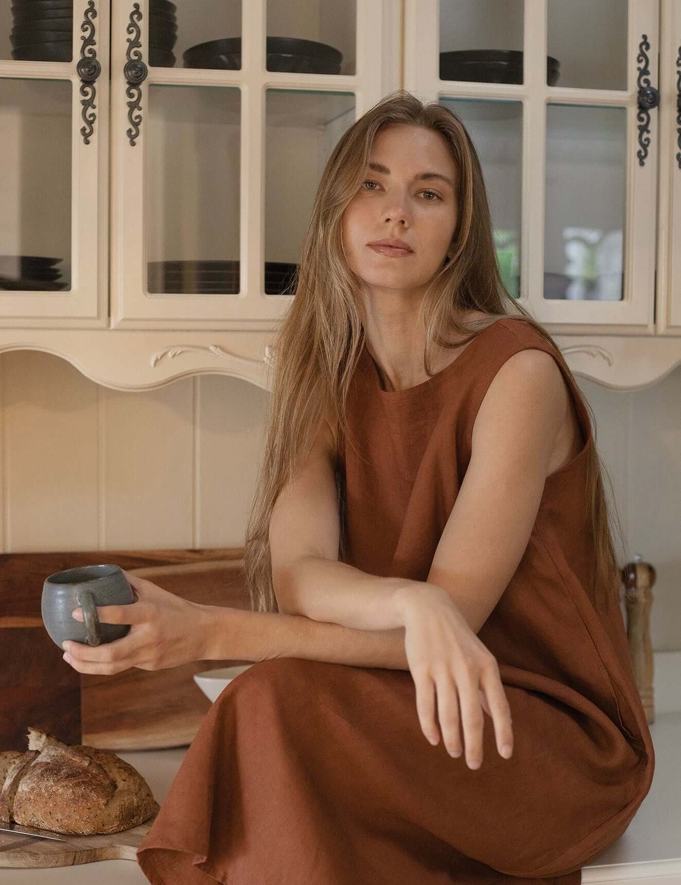 Woman wearing Cynthia Linen Dress Bark, sitting in a kitchen with a cup and bread, showcasing versatile style.
