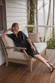 Charlotte Linen Dress Black worn by a model sitting on a couch, showcasing its effortless elegance and stylish design.