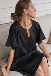 Charlotte Linen Dress Black worn by a model sitting on a couch, showcasing its effortless elegance and stylish design.