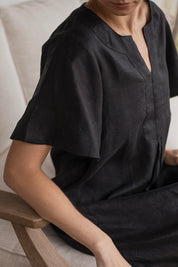 Charlotte Linen Dress Black worn by a model sitting on a couch, showcasing its effortless elegance and stylish design.