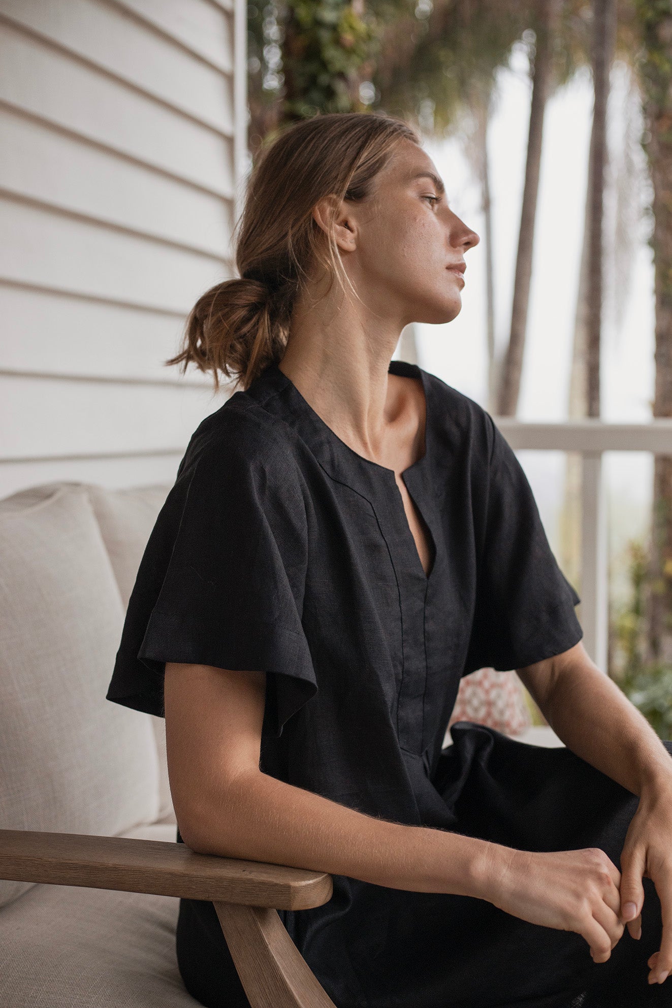 Charlotte Linen Dress Black worn by a model sitting on a couch, showcasing its effortless elegance and stylish design.