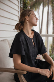 Charlotte Linen Dress Black worn by a model sitting on a couch, showcasing its effortless elegance and stylish design.