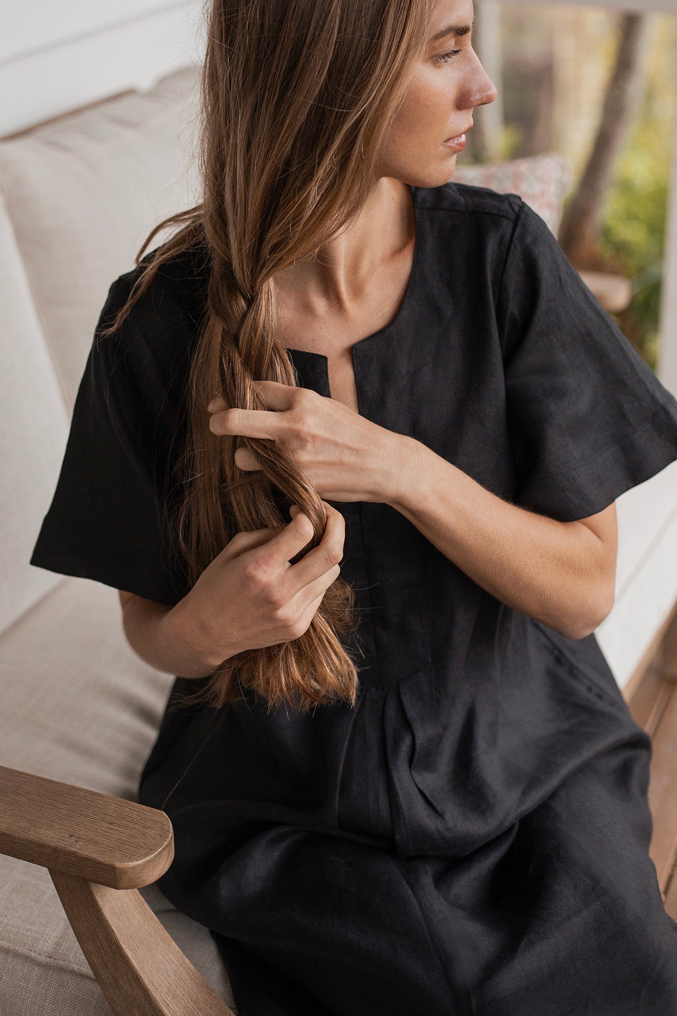 Charlotte Linen Dress Black worn by a model sitting on a couch, showcasing its effortless elegance and stylish design.