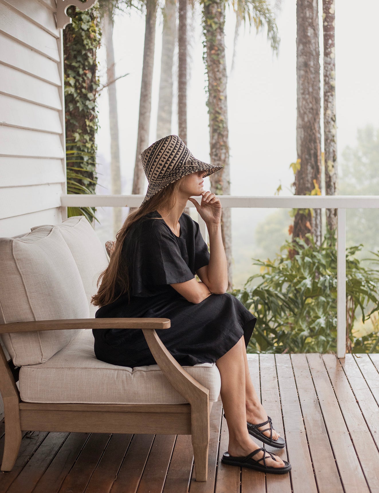 Charlotte Linen Dress Black worn by a model sitting on a couch, showcasing its effortless elegance and stylish design.