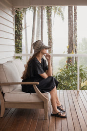 Charlotte Linen Dress Black worn by a model sitting on a couch, showcasing its effortless elegance and stylish design.
