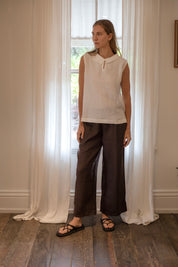 Model wearing Paris Linen Pants Cocoa paired with a white blouse, standing by a window with sheer curtains.