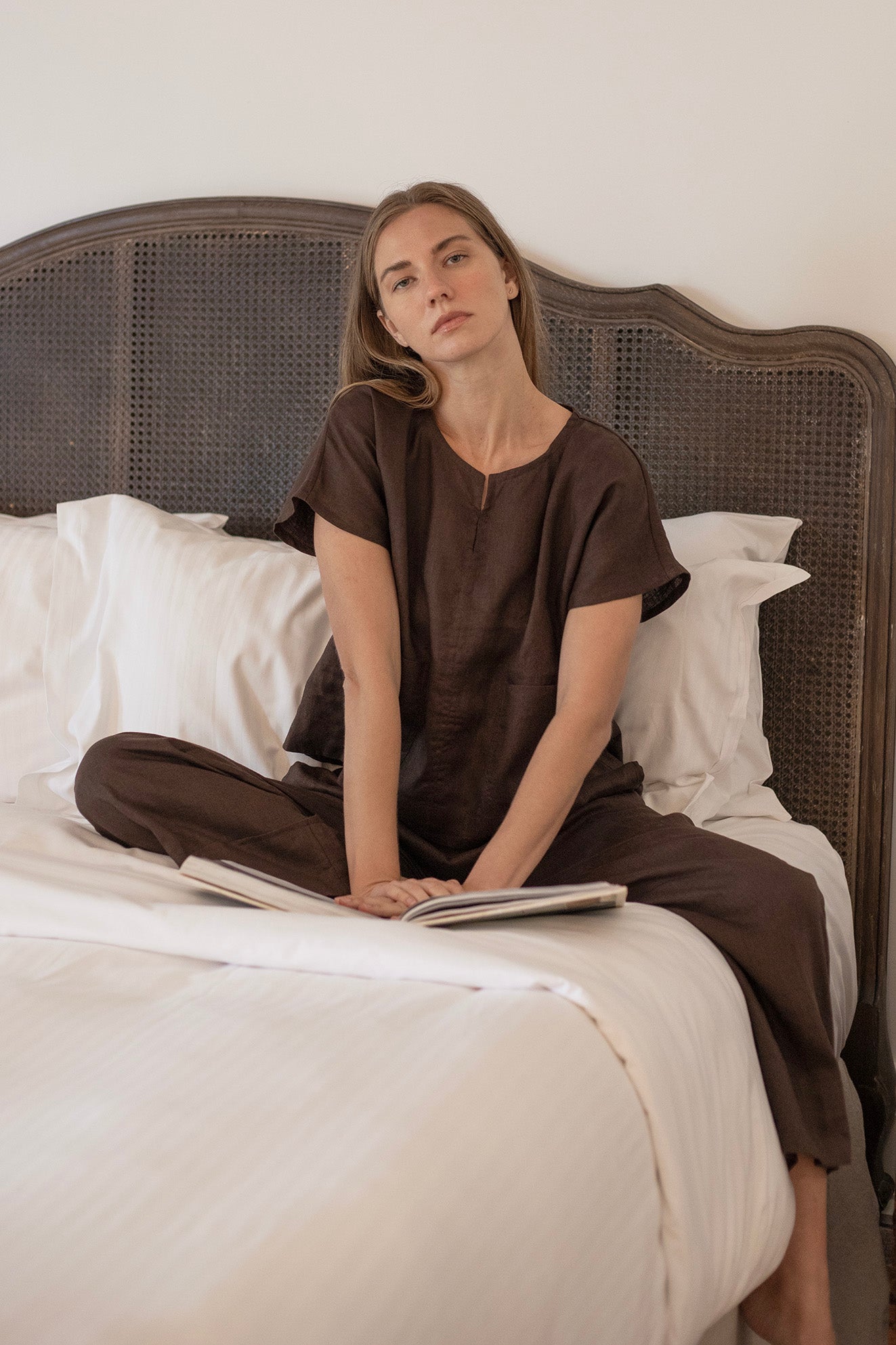 Model wearing Paris Linen Pants Cocoa seated on a bed, showcasing wide-leg design and comfortable style.
