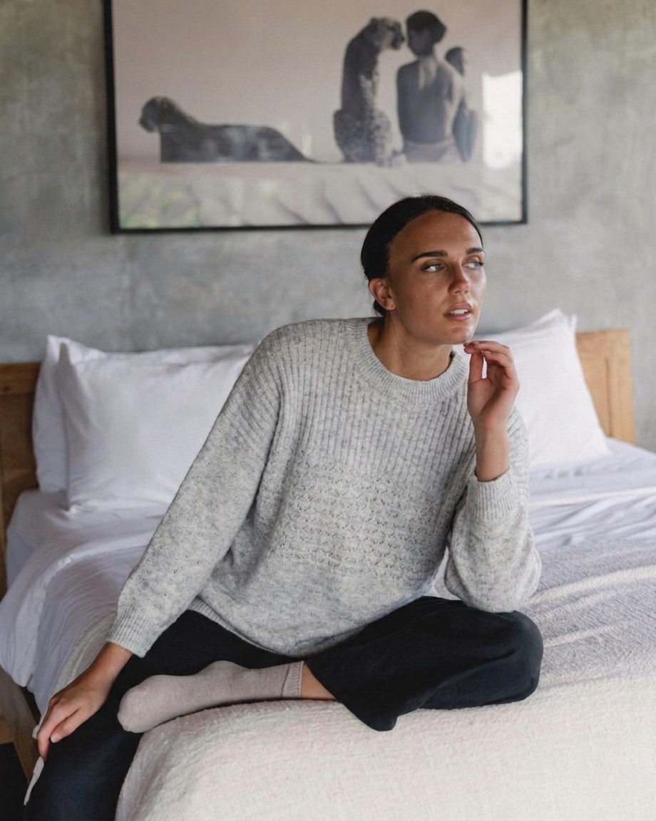 Woman sitting on a bed wearing the Marlo Wool Blend Jumper Dove Grey, showcasing its soft knit texture and relaxed fit.
