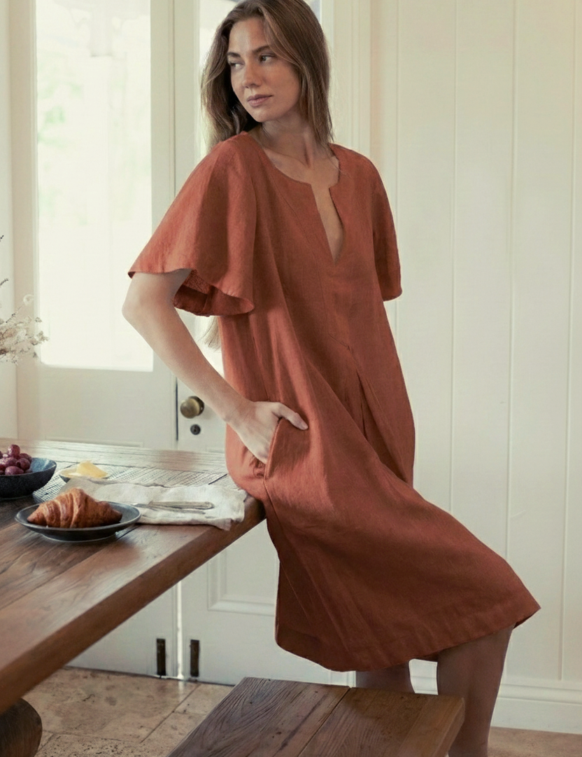 Model wearing Charlotte Linen Dress Bark while sitting at a table with food, showcasing effortless elegance with a sleeve.