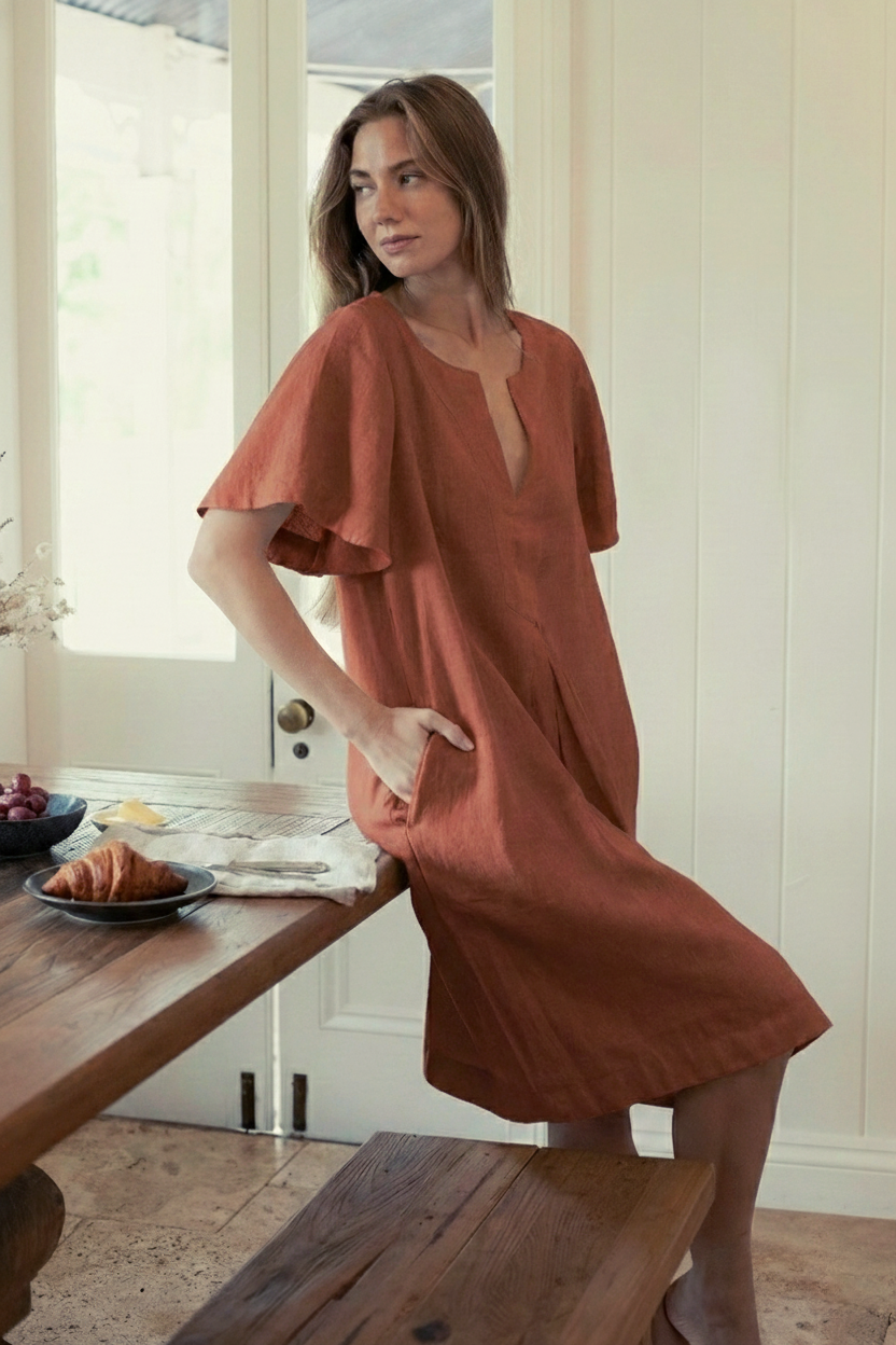 Model wearing Charlotte Linen Dress Bark while sitting at a table with food, showcasing effortless elegance with a sleeve.